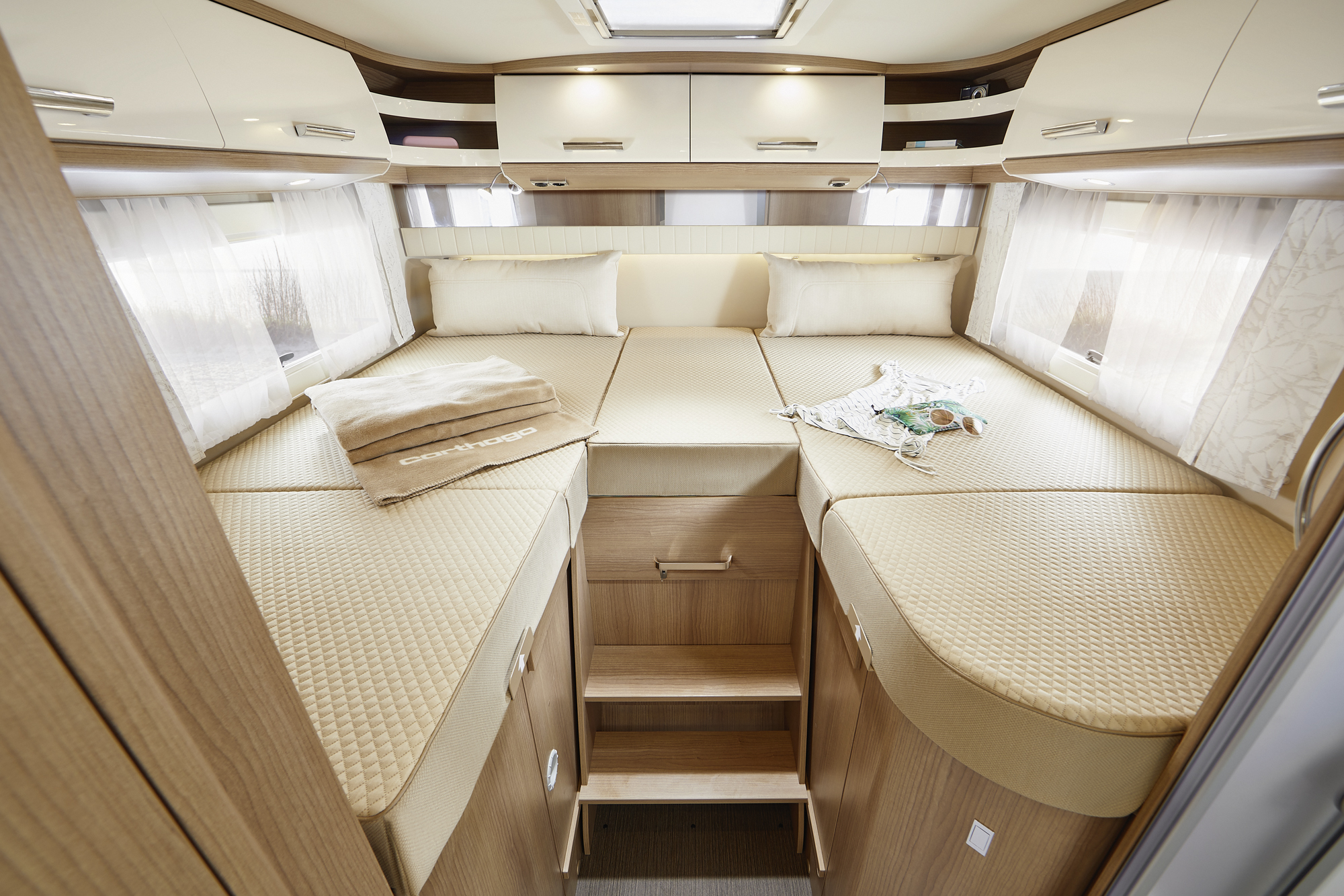 Modernes Wohnmobil-Schlafzimmer mit einem großen, ordentlich gemachten Bett, beiger Bettwäsche, Kissen, Hängeschränken und Handtüchern auf dem Bett. Stufen führen hinauf zum Schlafbereich, und natürliches Licht fällt durch ein Dachfenster ein.
