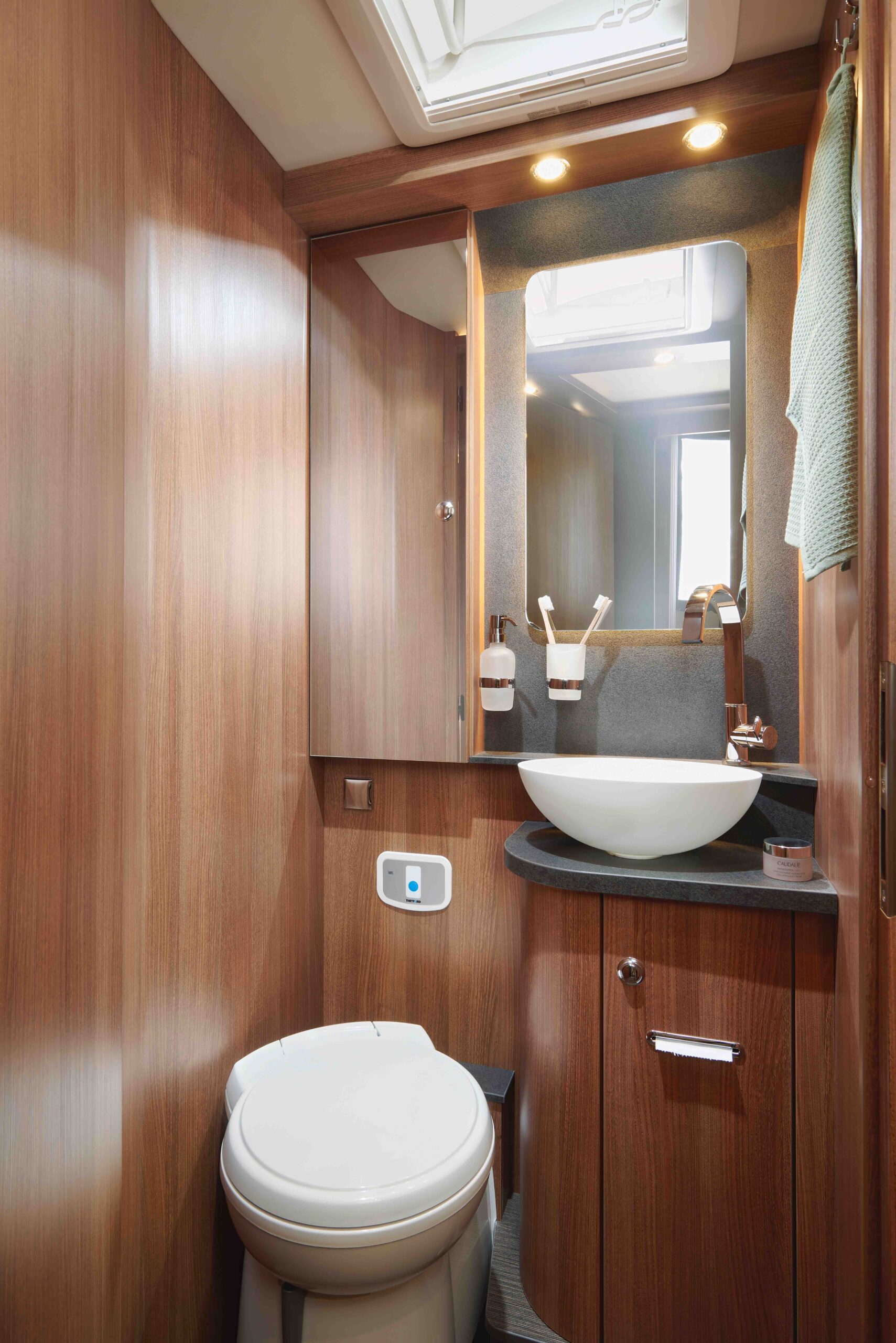 Kompaktes Badezimmer mit holzgetäfelten Wänden, einer runden weißen Toilette, einem modernen Waschbecken auf einer schwarzen Arbeitsplatte, einem Wandspiegel, Deckenleuchten und Schränken. Handtücher und Toilettenartikel sind übersichtlich angeordnet.