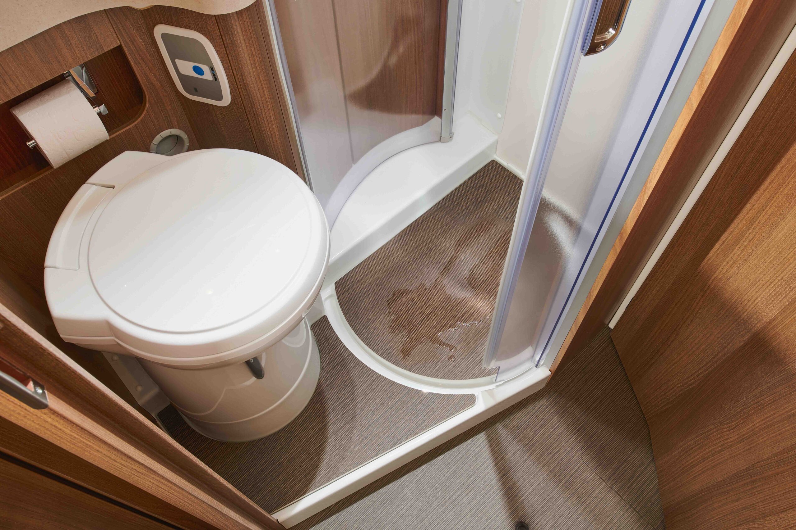 Ein kompaktes Badezimmer mit einer weißen Toilette neben einer geschwungenen Duschkabine, braunen holzgetäfelten Wänden und einem nassen Fußboden in der Nähe des Duscheinstiegs. Ein Toilettenpapierhalter ist an der Wand angebracht.