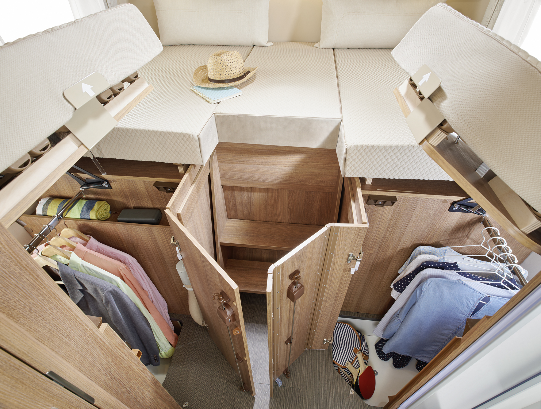 Das Bild zeigt ein organisiertes Schranksystem unter einem Hochbett mit offenen Holzschränken, in denen hängende Kleidung, gefaltete Kleidungsstücke und ein Hut auf dem Bett darüber aufbewahrt werden. Der Raum ist hell und übersichtlich gestaltet.