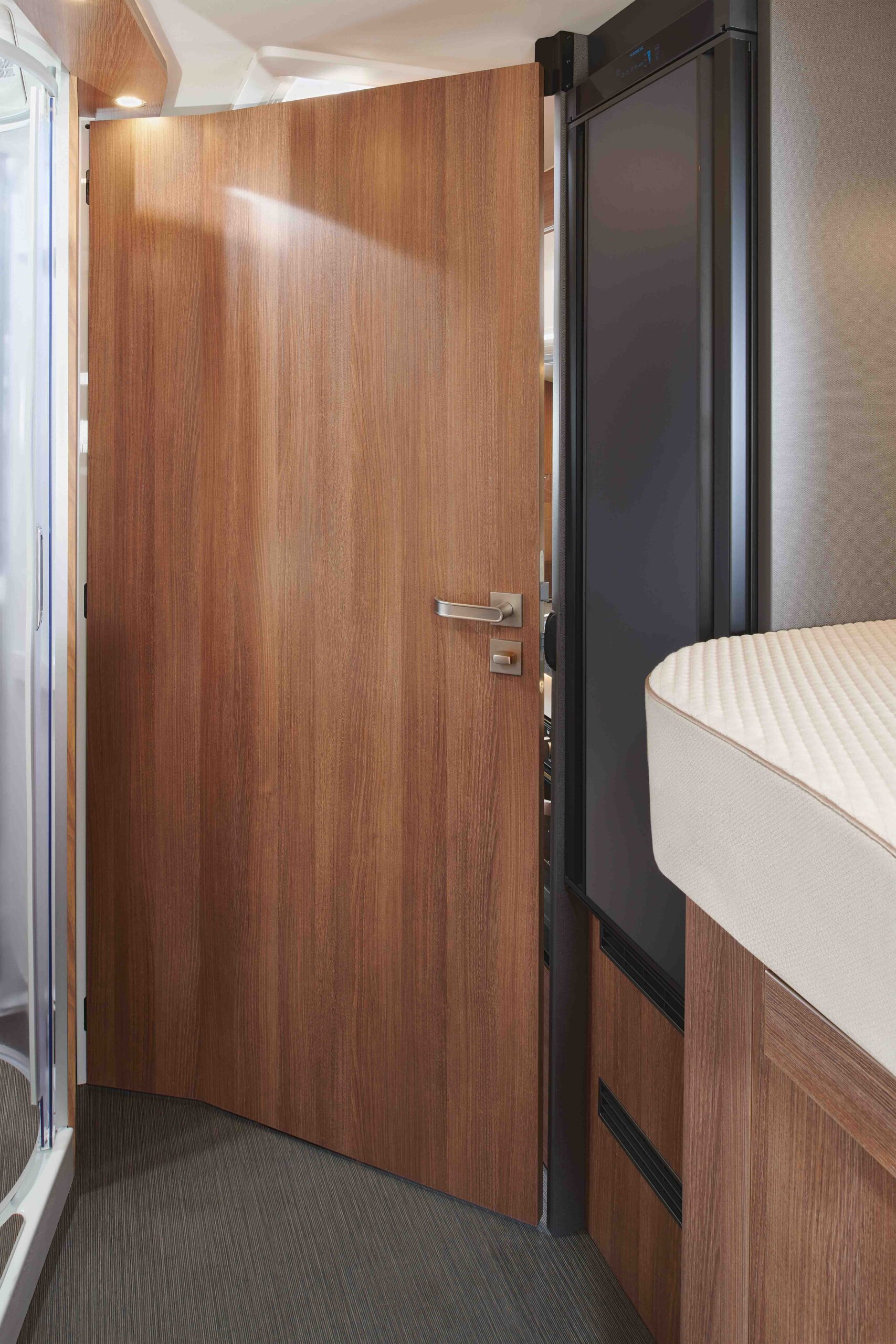 Eine Holztür mit modernen silbernen Griffen ist in einem kompakten, zeitgenössischen Interieur teilweise geöffnet und gibt den Blick auf einen Teil eines Bettes mit einer beigen Matratze und einem schwarzen Gerät neben der Tür frei.