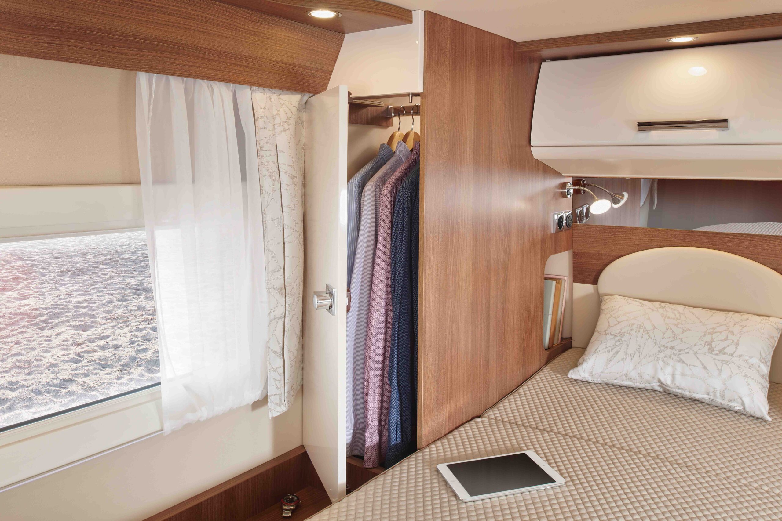 Ein gemütliches Camper-Schlafzimmer mit Bett, Kissen und Tablet, Holzschränken, einem kleinen Kleiderschrank mit aufgehängter Kleidung, einem Fenster mit Vorhängen und sanfter Beleuchtung, die eine warme Atmosphäre schafft.