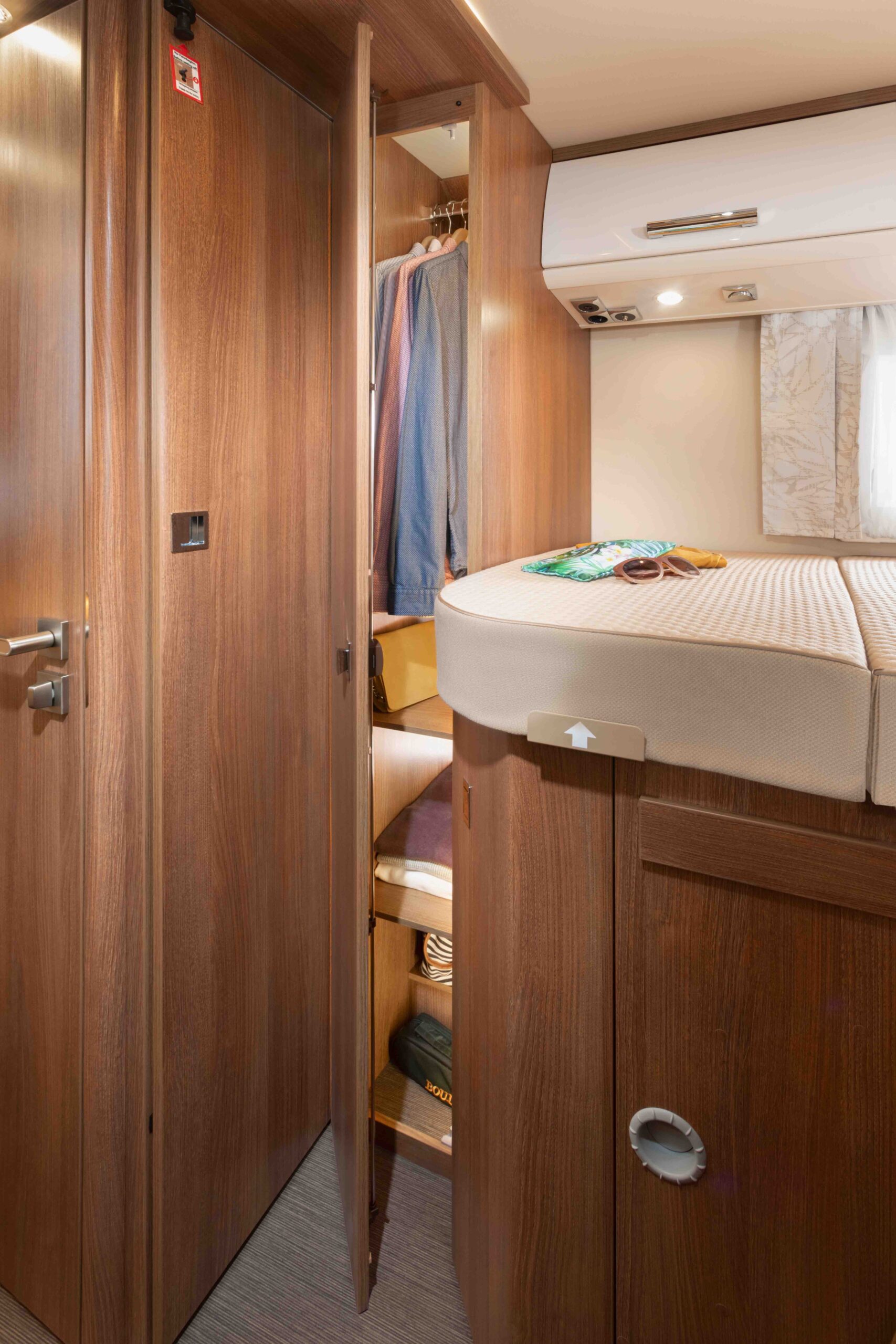 In einem kompakten Schlafzimmer steht ein erhöhtes Bett mit einer Matratze und Büchern darauf, daneben ein teilweise offener Holzschrank mit Kleiderbügeln und Regalen. Die holzgetäfelten Wände und Schränke werden durch warmes Licht hervorgehoben.