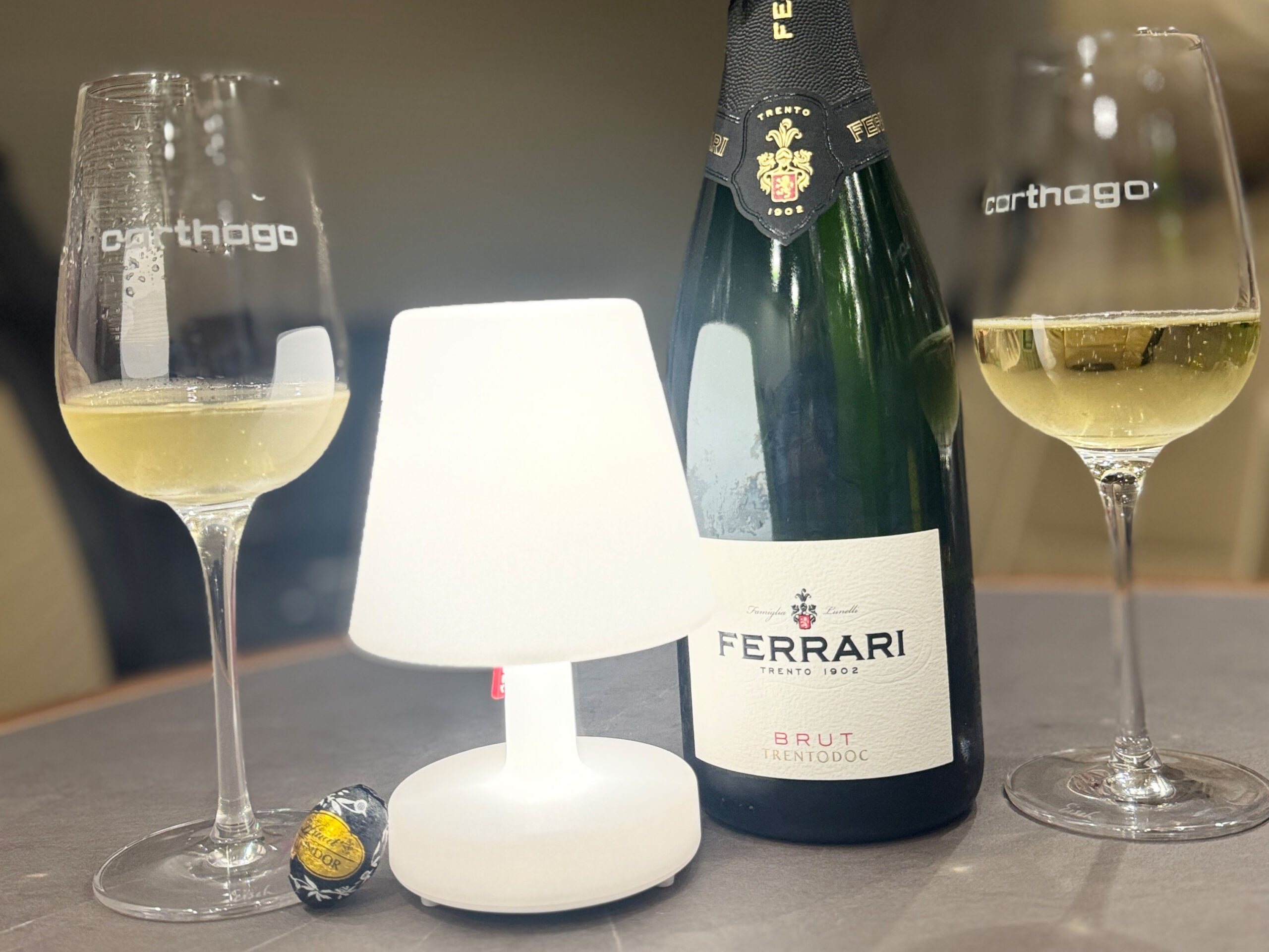 Accanto a due bicchieri con il logo Carthago c'è una bottiglia di Ferrari Brut. Al centro brilla una lampada da tavolo, accanto c'è una pralina ancora nella confezione: tutto questo crea un'atmosfera accogliente, perfetta per condividere le esperienze di viaggio, come un resoconto di un viaggio in Finlandia.