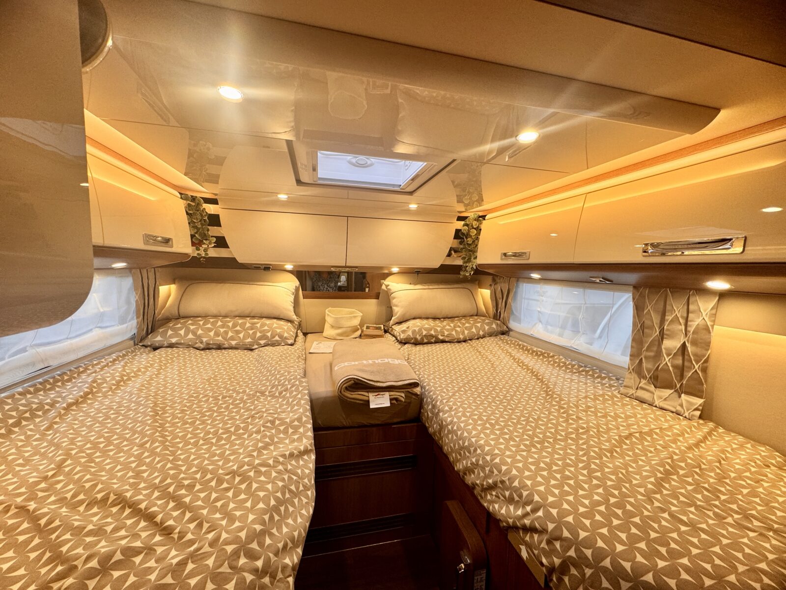 Due letti singoli con lenzuola a motivi geometrici in un'accogliente e luminosa camera da letto del camper: l'ideale per rilassarsi dopo una lunga giornata trascorsa a scrivere il tuo diario di viaggio sulla Finlandia. Gli armadietti pensili, le lampade da lettura e le finestre laterali con tende creano un ambiente accogliente e ordinato.