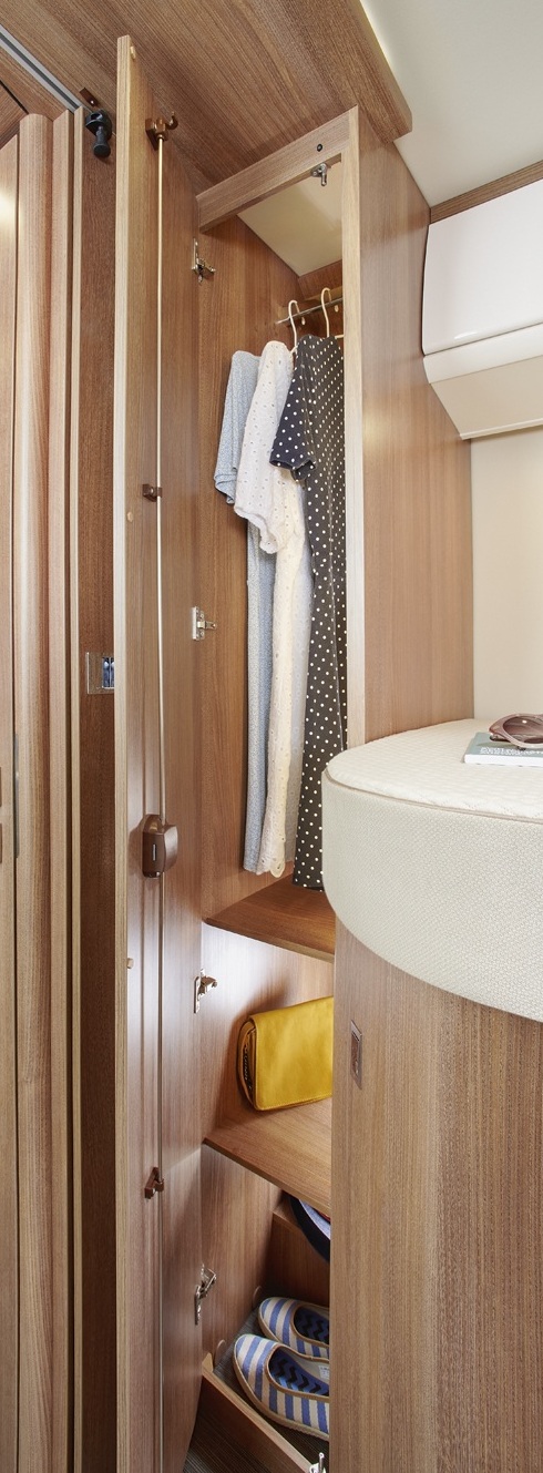Ein offener Kleiderschrank aus Holz, in dem ein blaues Kleid und ein gepunktetes Kleid hängen; auf den Regalen darunter liegen eine gelbe Tasche und gestreifte Handtücher; daneben steht ein geschwungenes Bett mit einem beigen Kissen.