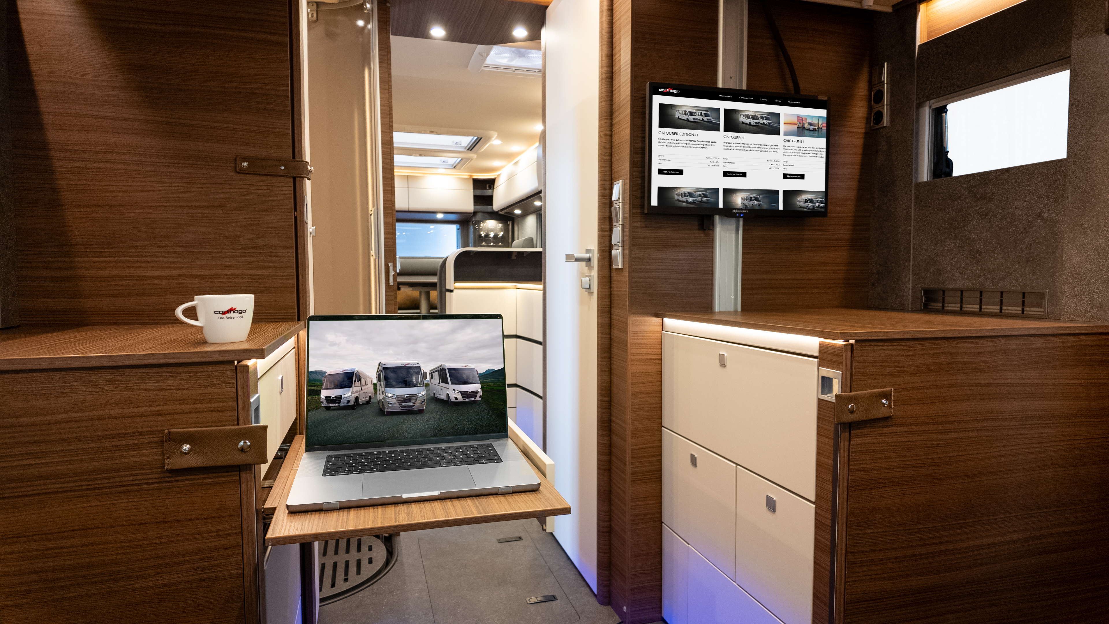 Ein modernes Wohnmobil-Interieur mit Holzakzenten, einem ausziehbaren Schreibtisch, auf dem ein Laptop mit Bildern von Wohnmobilen steht, einem an der Wand montierten Monitor, auf dem eine Website angezeigt wird, und einer Kaffeetasse auf einem Schrank.