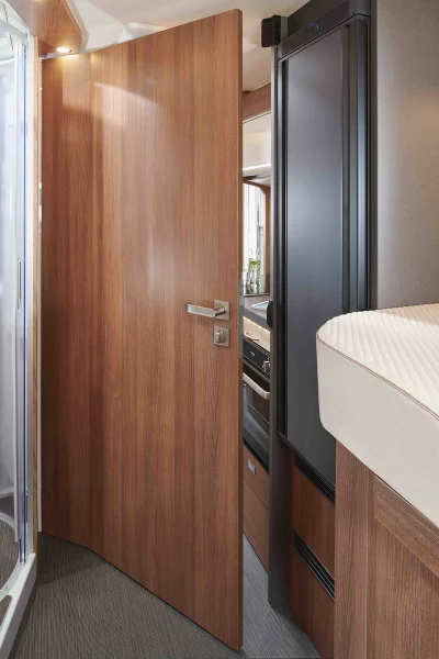 Eine halb geöffnete Holztür gibt den Blick frei auf den kompakten Innenraum eines Carthago C2-Tourer-Wohnmobils mit Kühlschrank, Kochnische und einem Hochbett mit beiger Matratze.