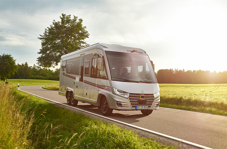 Ein weißes Wohnmobil fährt auf einer kurvenreichen Landstraße, die von grünem Gras und Bäumen gesäumt ist, während im Hintergrund die Sonne scheint.