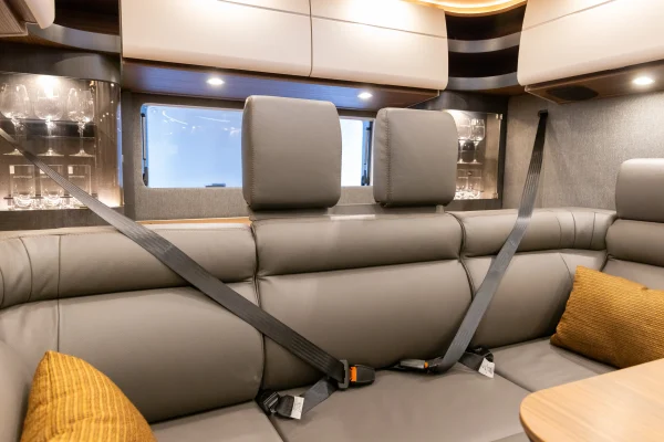 Moderne Wohnmobil-Innenausstattung, inspiriert vom Carthago Liner for Two, mit einer grauen Ledersitzbank mit angeschnallten Sicherheitsgurten, zwei senfgelben Kissen, Glasschränken und Stauraum an der Decke. Ein Fenster über dem Sofa lässt Tageslicht herein.