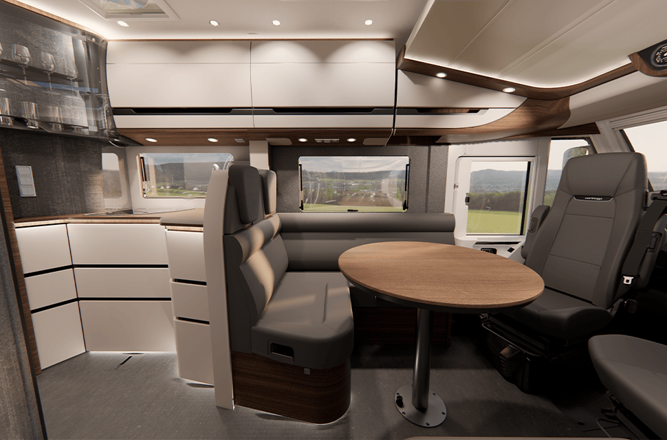Moderner Wohnmobil-Innenraum mit grauen Sitzen, einem runden Holztisch, weißen Schränken und großen Fenstern, die den Blick nach draußen freigeben. Der Raum wirkt aufgeräumt, hell und ist auf Komfort und Funktionalität ausgelegt.