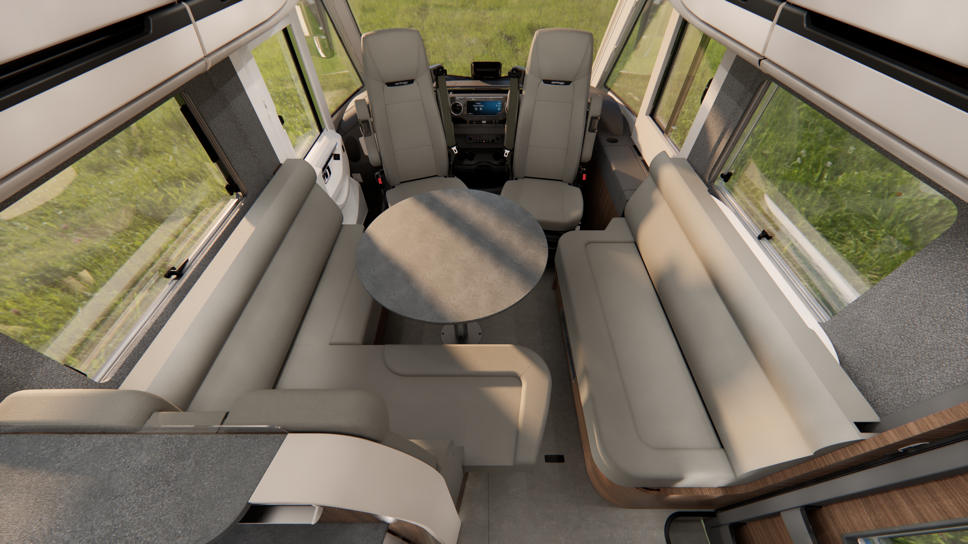 Innenansicht eines Wohnmobils mit zwei Vordersitzen, einem runden Tisch in der Mitte und beigen Sofas, die in U-Form angeordnet sind. Große Fenster sorgen für Tageslicht und bieten einen Blick auf die Wiese draußen.