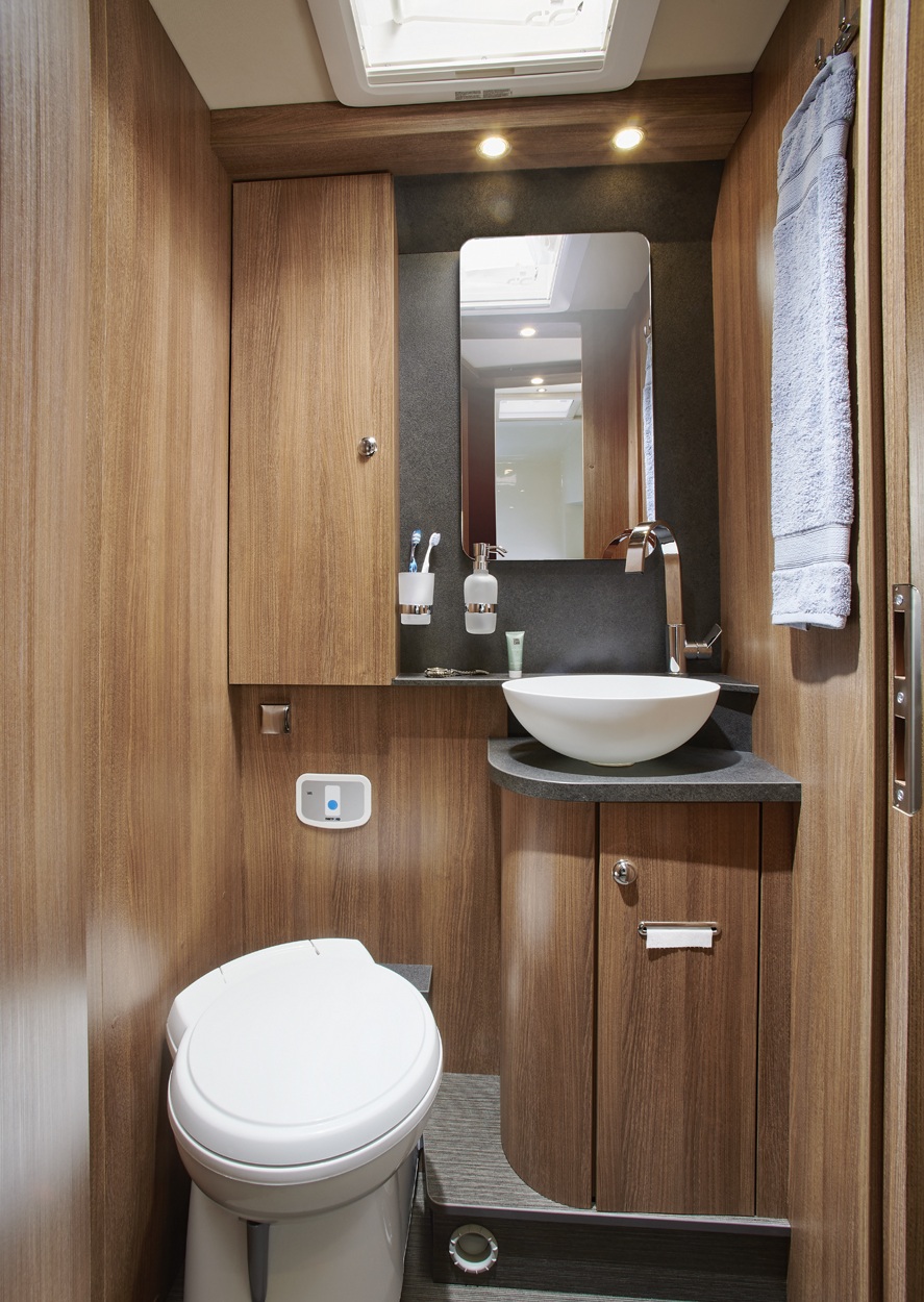 Kleines Badezimmer mit Holzvertäfelung, einer weißen Toilette, einem Waschbecken mit moderner Armatur, einem Spiegel, Pflegeprodukten, einem Handtuch auf einem Handtuchhalter, Schränken und Deckenleuchten. An der Decke ist ein Oberlicht zu sehen.