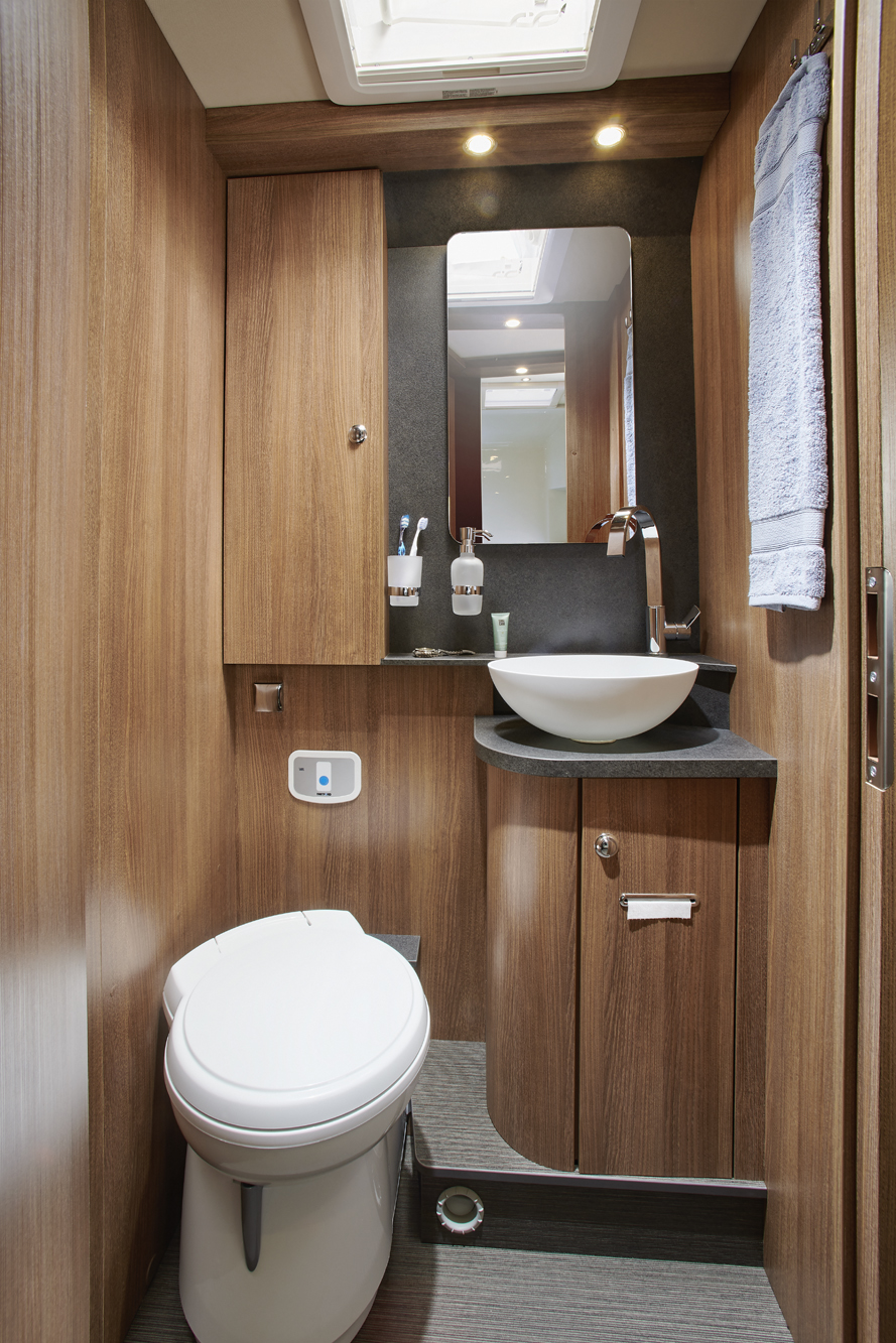 Kompaktes Badezimmer mit Holzvertäfelung, einer weißen Toilette, einem Waschbecken mit Aufsatzwaschschüssel, einem an der Wand montierten Spiegel, einem Handtuch und Pflegeprodukten. Ein Oberlicht sorgt für Tageslicht. Modernes und effizientes Design für Wohnmobile oder kleine Räume.
