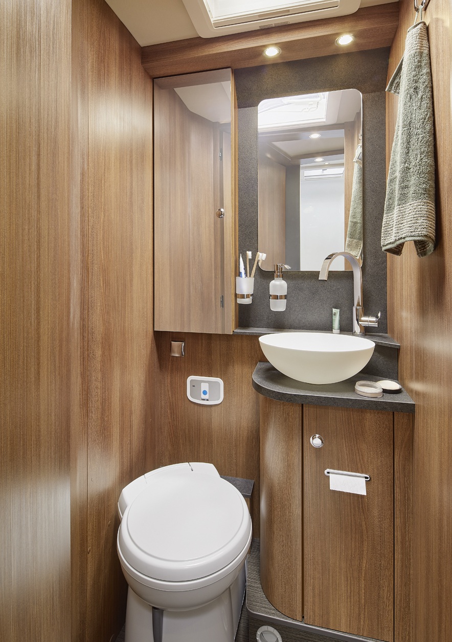 Kompaktes Badezimmer mit Holzvertäfelung, einer runden weißen Toilette, einem Aufsatzwaschbecken, einem Spiegel, einem Handtuch und Pflegeprodukten auf einer schwarzen Waschtischplatte. Deckenleuchten erhellen den Raum.