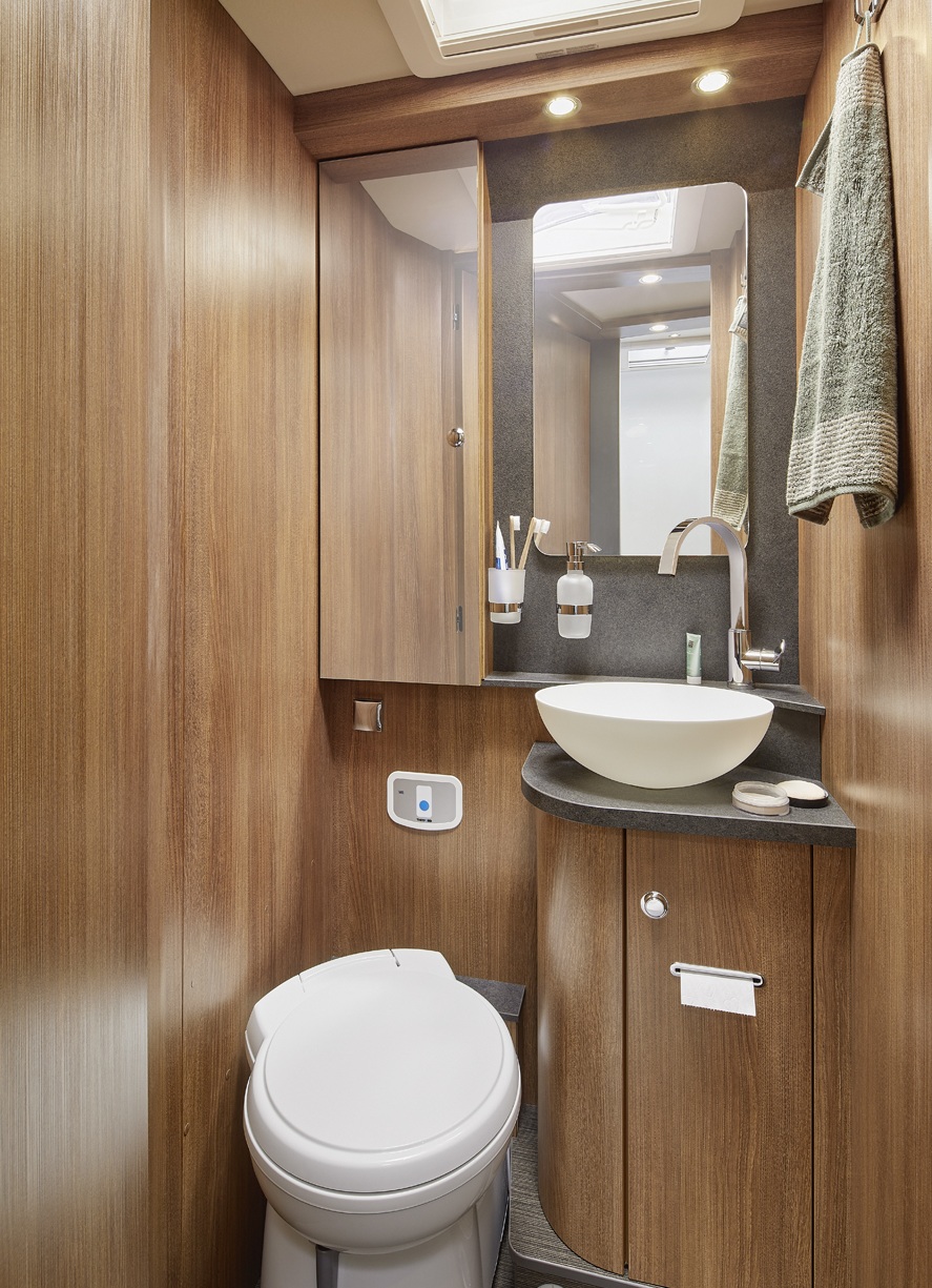 Kleines Badezimmer mit Holzvertäfelung, einem runden weißen Waschbecken auf einer schwarzen Arbeitsplatte, einem Spiegel, einer Toilette, Handtüchern, Toilettenartikeln und Deckenleuchten. Modernes, kompaktes Design.
