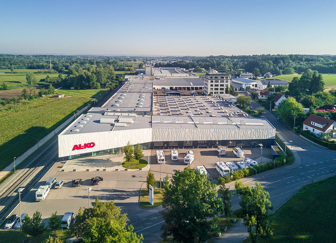 Luftaufnahme eines großen Industriegebäudes mit dem AL-KO-Logo in der Nähe von Carthago City in Aulendorf, umgeben von Bäumen, geparkten Fahrzeugen und grünen Feldern an einem sonnigen Tag. In der Nähe sind Wohnhäuser und Straßen zu sehen.