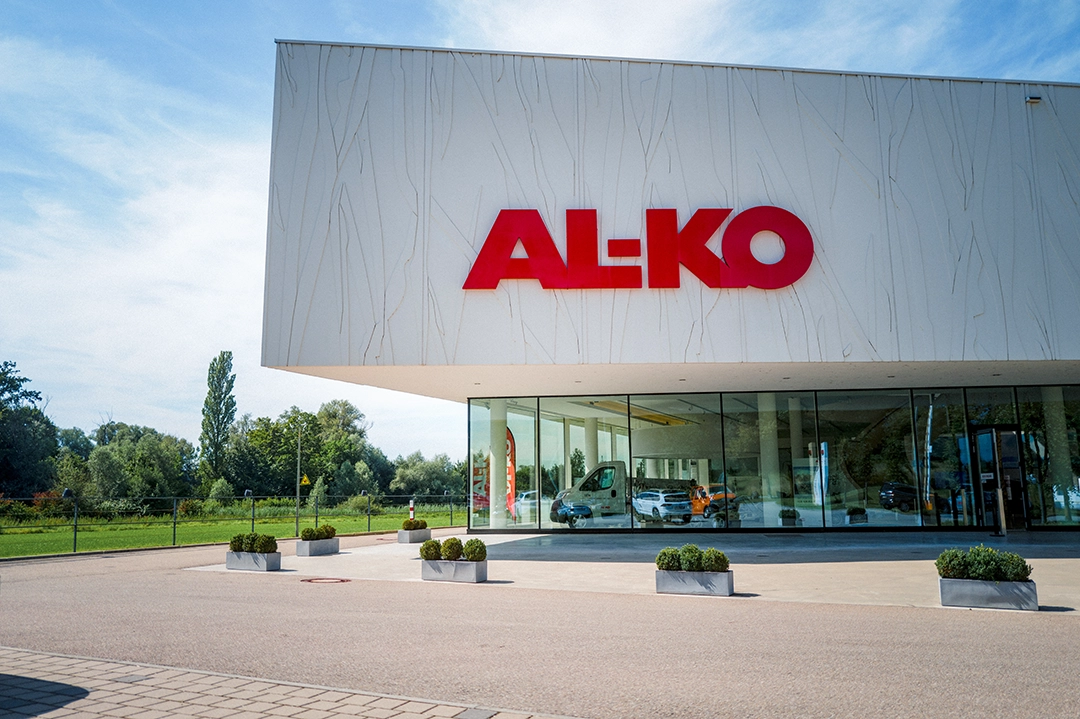 Ein modernes Gebäude mit den großen roten Buchstaben „AL-KO“ an der Fassade, mit Glaswänden und Topfpflanzen vor dem Eingang – Carthago City in Aulendorf ist das Ziel, umgeben von grünen Bäumen und Rasenflächen im Hintergrund.