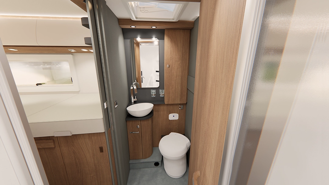 Kompaktes Wohnmobil-Badezimmer im Carthago C2-Tourer mit einer runden weißen Toilette, einem kleinen Waschbecken, einem Spiegel, Holzschränken und einer Milchglastür. Links ist der Schlafbereich mit einem Bett zu sehen. Deckenleuchten und ein Dachfenster sorgen für Helligkeit im Raum.