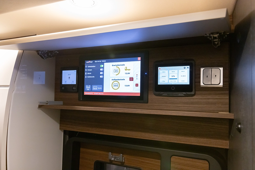 Bedienfeld mit drei digitalen Displays und zwei Schaltern an einer Holzwand, auf dem verschiedene Einstellungen und Energieinformationen angezeigt werden, vermutlich im Inneren eines Carthago Chic E-Line-Wohnmobils.