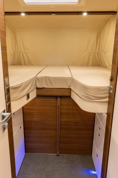 Ein kompakter Raum mit einem erhöhten Doppelbett, beiger Bettwäsche, Holzwänden, weißen Schränken darunter und einem Oberlicht darüber. Der Raum scheint sich im Inneren eines „Carthago Liner for Two“-Wohnmobils zu befinden.