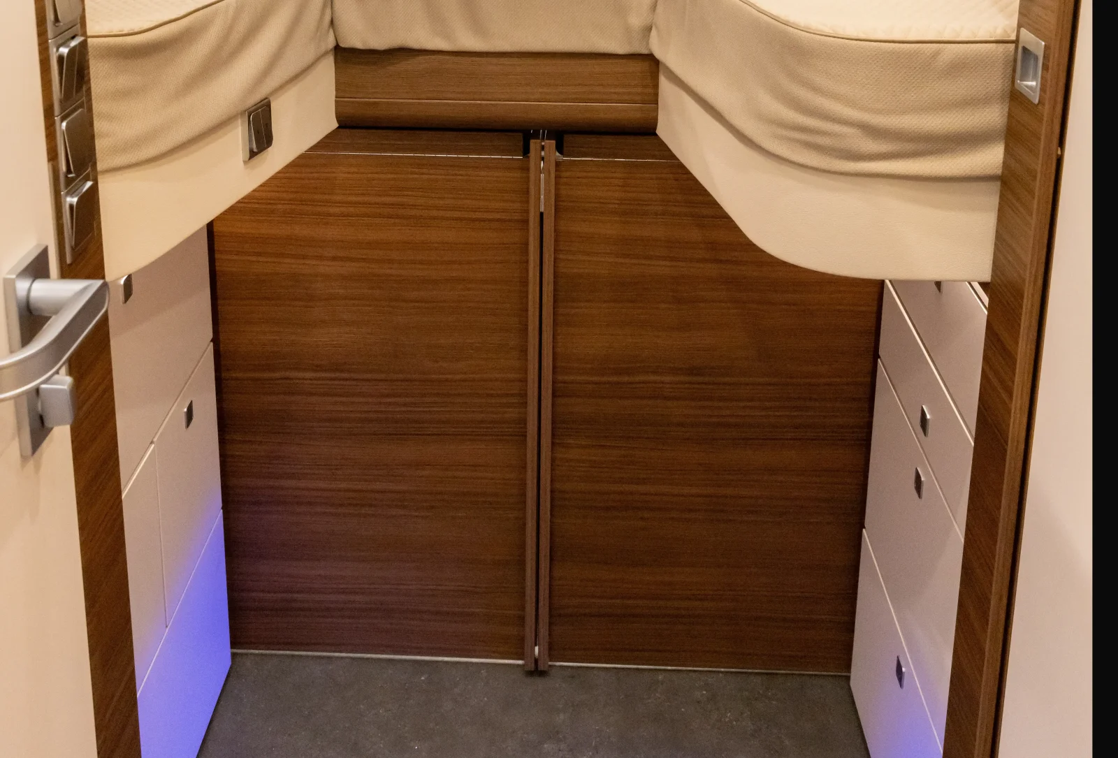 Ein kleiner Raum aus Holz im Inneren des Carthago Liner For Two verfügt über ein Hochbett, Stauraumschubladen auf beiden Seiten und eine sanfte violette Beleuchtung in Bodennähe. Auf der linken Seite ist ein Türgriff zu sehen.