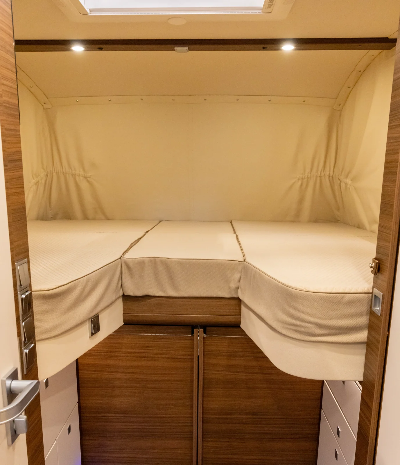 Ein kompaktes Schlafzimmer im Carthago Liner for Two verfügt über ein ordentlich gemachtes Bett mit cremefarbener Bettwäsche, darunter Schränke mit Holzverkleidung sowie beidseitig eingebaute Schubladen. Der Raum wird durch sanfte Beleuchtung und eine beige Stofftapete aufgewertet, wodurch eine gemütliche und einladende Atmosphäre entsteht.