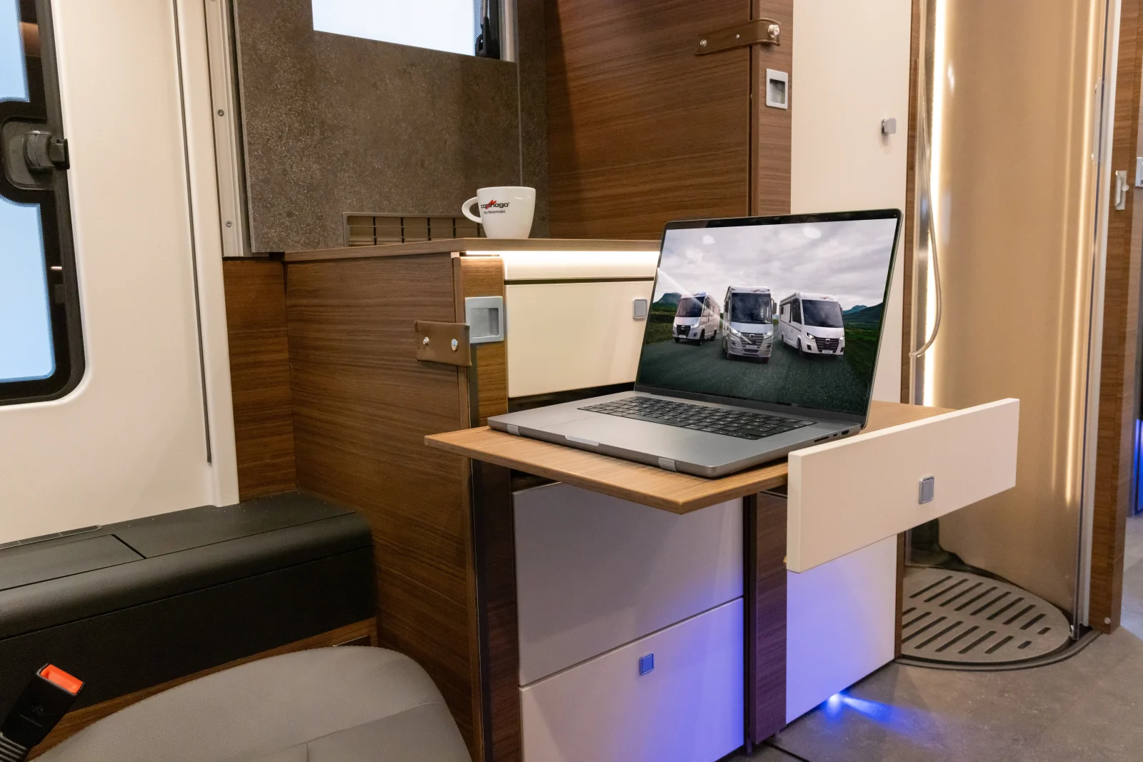 In einem modernen Carthago Liner for Two-Wohnmobil steht ein Laptop, auf dessen Bildschirm Bilder von Wohnmobilen zu sehen sind, auf einem ausziehbaren Holzregal; auf der Arbeitsplatte steht eine Tasse, und der mit Holz vertäfelte Innenraum ist zu sehen.