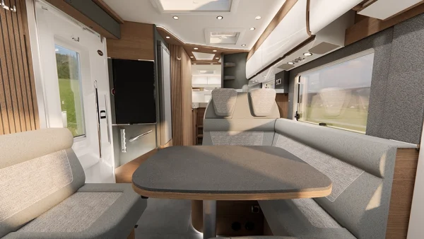 Moderne Wohnmobil-Innenausstattung des Carthago C2-Tourer mit hellen Holzakzenten, grauen Polstersitzen rund um einen Esstisch, großen Fenstern, einem Fernseher und einem kompakten Küchenbereich im Hintergrund. Hell und geräumig mit viel Tageslicht.