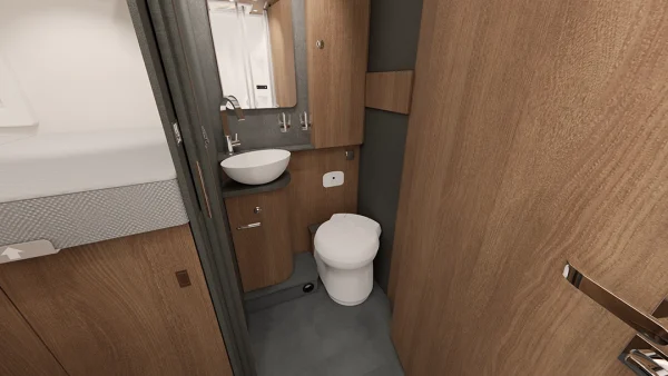Ein kompaktes Badezimmer mit Holzschränken, einem runden weißen Waschbecken, einem kleinen Spiegel und einer weißen Toilette neben einem Bett. Der Raum scheint sich im Inneren eines Carthago C2-Tourer-Wohnmobils oder eines Tiny House zu befinden.
