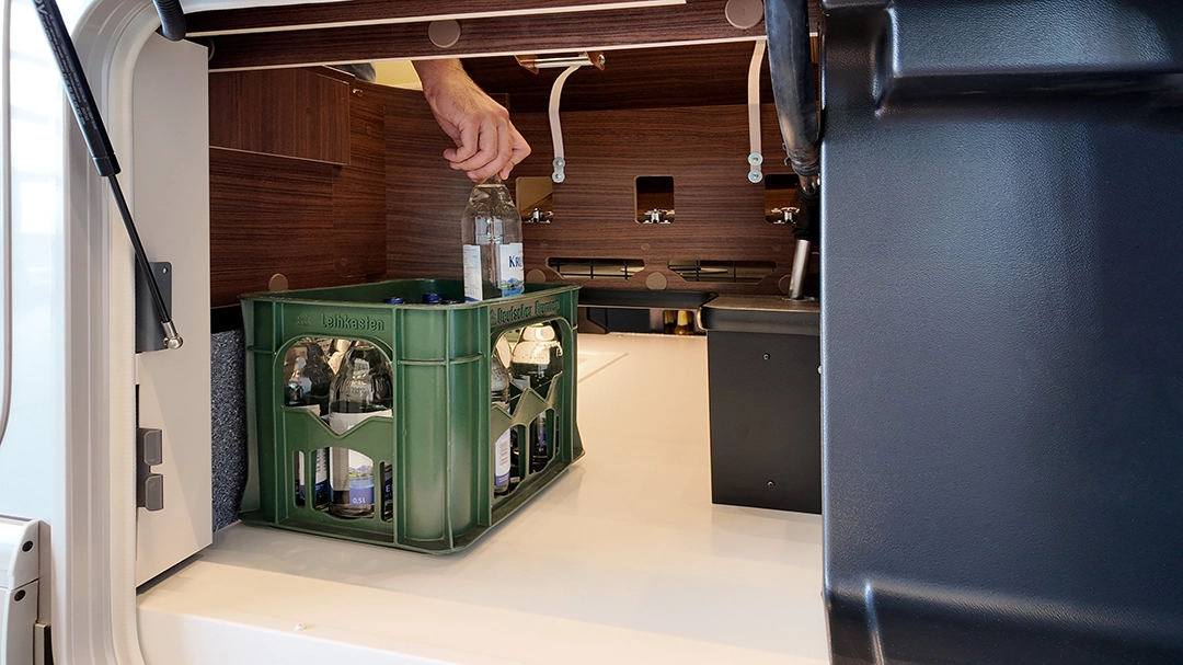 Eine Hand legt eine Flasche in eine grüne Kiste, die bereits mit Flaschen gefüllt ist und sich im Stauraum eines Wohnmobils – genauer gesagt des Carthago Chic E Line – befindet, wobei die Holzverkleidung und die Wasserleitungen zu sehen sind.