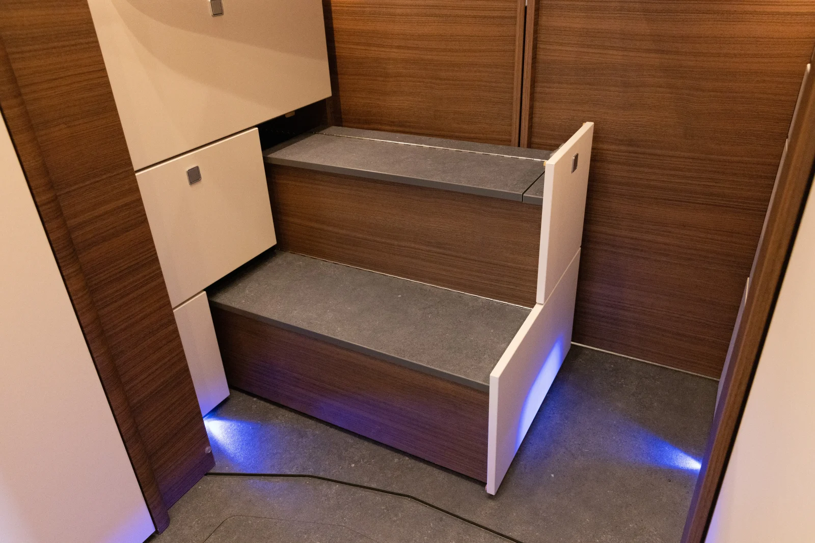 Moderne Holzstufen mit weißen Seitenwänden und eingebauten Schubladen, die von blauen LED-Leuchten an der Unterseite beleuchtet werden, eingebettet in den kompakten, modernen Innenraum des Carthago Liner for Two.