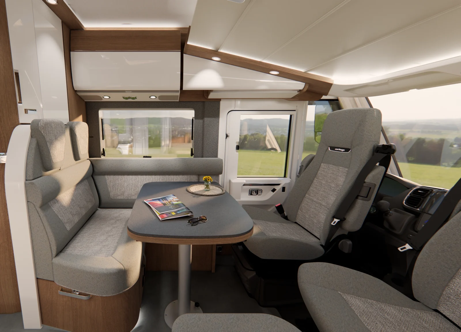 Innenraum eines Carthago C2-Tourer-Wohnmobils mit einem Tisch, zwei Sitzbänken und zwei Vordersitzen. Auf dem Tisch liegen Zeitschriften, ein Schlüssel und eine kleine Blumenvase. Durch die großen Fenster ist draußen eine grasbewachsene Landschaft zu sehen.