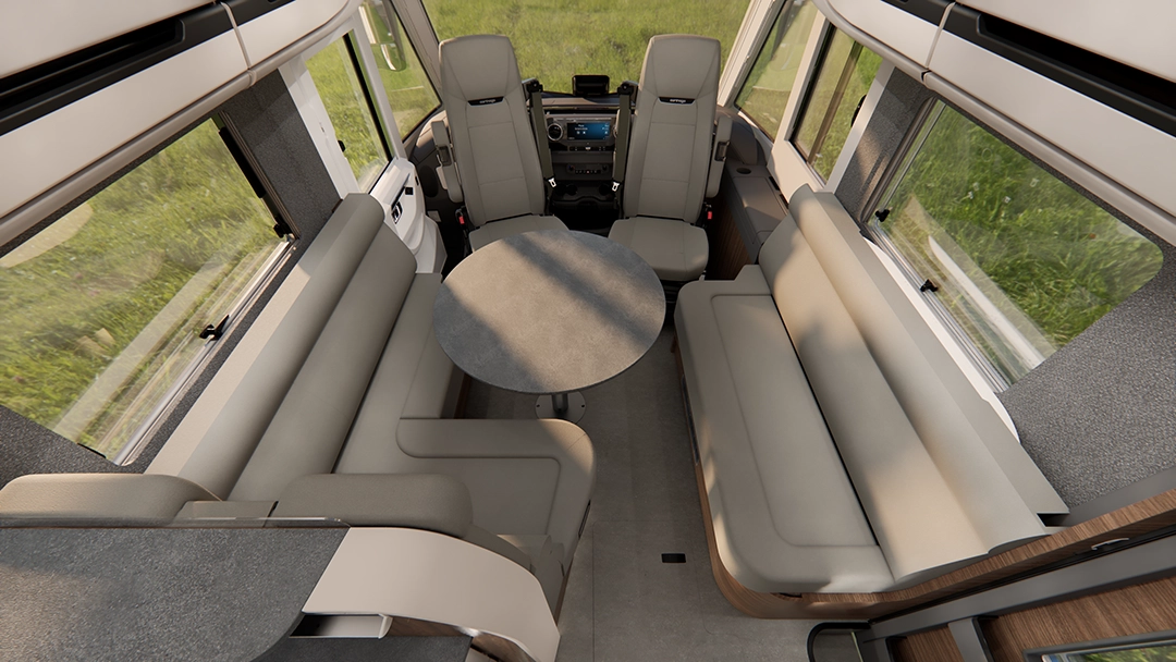 Innenraum eines Wohnmobils der Carthago Chic E-Line mit zwei langen beigen Sofas, einem runden Tisch in der Mitte, großen Fenstern und dem vorne sichtbaren Fahrerbereich.