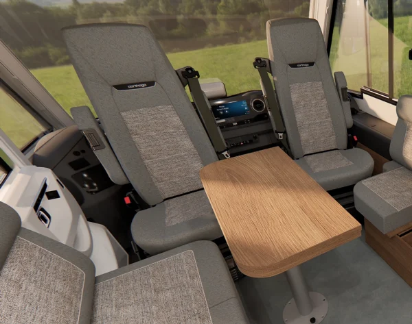Ein modernes Wohnmobil-Interieur mit zwei grauen Kapitänsstühlen, die einem Holztisch zugewandt sind, großen Fenstern, die den Blick auf die üppige grüne Landschaft draußen freigeben, und einem Armaturenbrett mit Touchscreen-Display – inspiriert vom raffinierten Design und Komfort des Carthago c2-Tourer.
