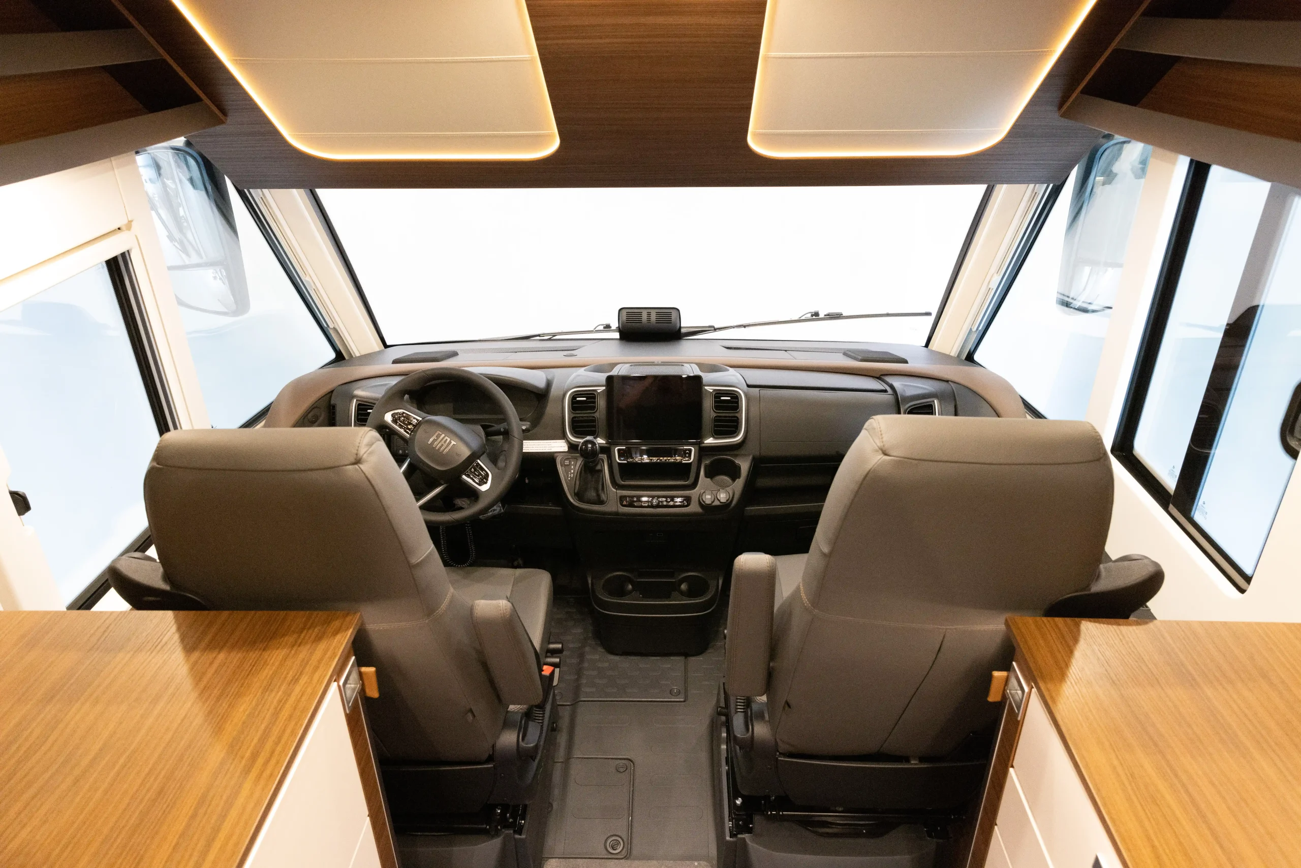 Blick auf das Cockpit eines modernen Carthago Liner for Two-Wohnmobils mit zwei grauen Sitzen, einem Lenkrad, einem zentralen Touchscreen-Display und Akzenten aus Holz, vom Wohnbereich aus in Fahrtrichtung gesehen.