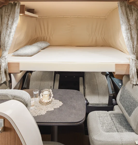 Innenraum eines Carthago C2-Tourer-Wohnmobils mit einem Hochbett über einem Tisch und einer Sitzecke. Das Bett ist mit einem Kissen ausgestattet und durch Vorhänge abgetrennt. Auf dem Tisch darunter stehen Kerzen und liegt eine dekorative Spitzendecke, während gepolsterte Sitze den Tisch umgeben.
