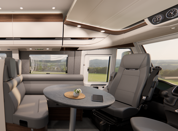 Ein modernes Wohnmobil-Interieur, inspiriert von der Carthago Chic E-Linie, mit grauen Sitzgelegenheiten, einem runden Tisch, auf dem eine kleine Topfpflanze und ein Smartphone stehen, großen Fenstern, Hängeschränken und einem Blick auf grüne Felder draußen.
