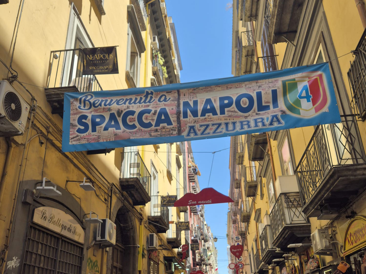 Un grande striscione con la scritta «Benvenuti a Spacca Napoli Azzurra» è appeso sopra una stradina di Napoli, in Italia, fiancheggiata da edifici gialli e balconi, che offre un quadro vivace, ben diverso da qualsiasi resoconto di viaggio dalla Finlandia. Negozi e piccole insegne costeggiano la strada affollata.