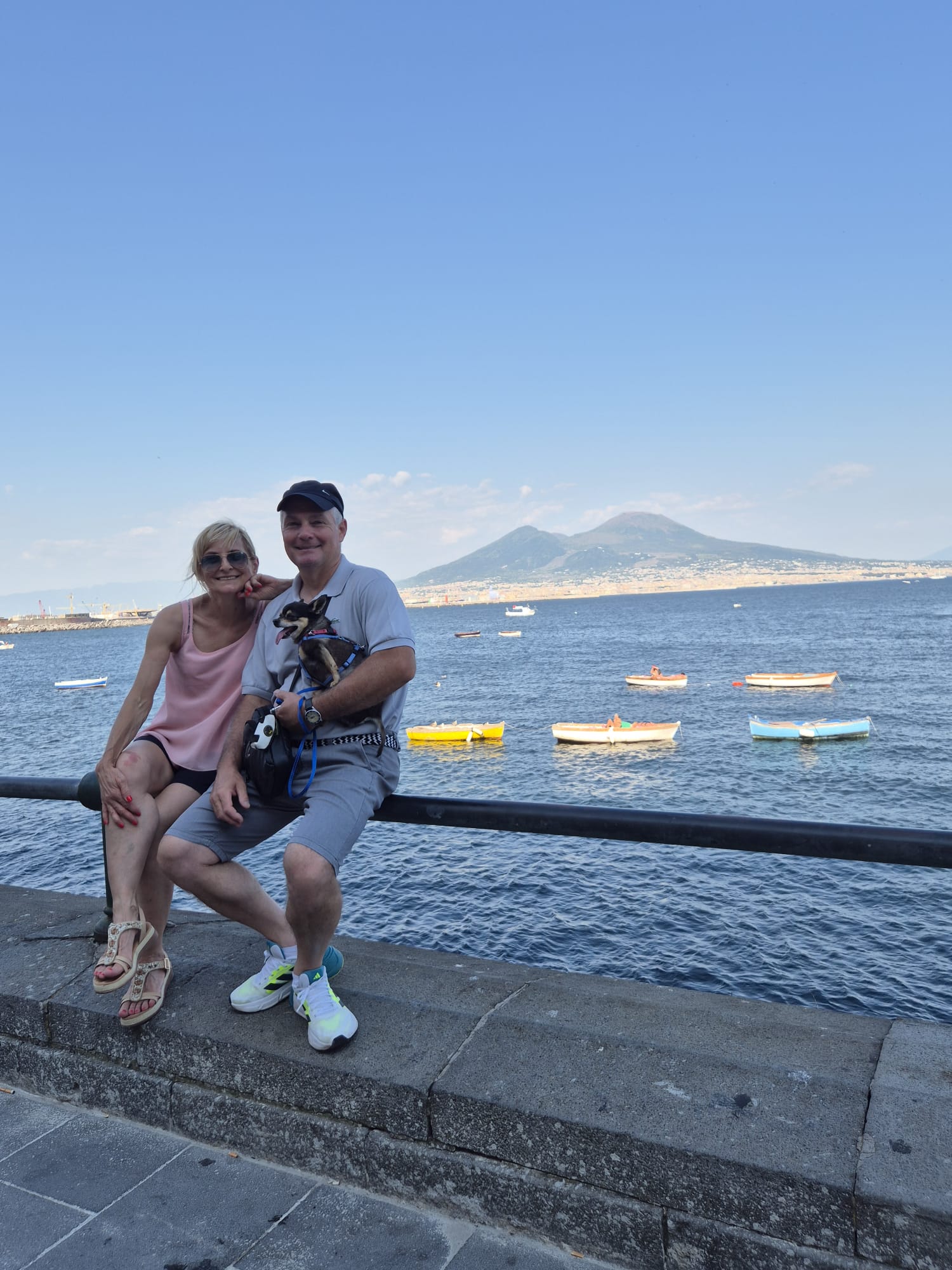 Una coppia sorridente con un cagnolino è seduta su un muretto di pietra in riva all'acqua. Barche colorate galleggiano sul mare e, in lontananza, una montagna si staglia contro il cielo di un azzurro splendente: lo scenario perfetto per un’avventura da raccontare in un diario di viaggio sulla Finlandia.