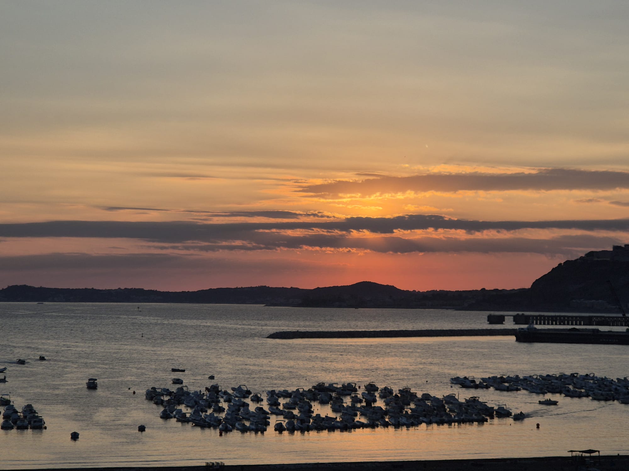 Bei Sonnenuntergang liegen Boote in einem ruhigen Hafen vor Anker, während vereinzelte Wolken den orange-rosa Himmel teilweise verdecken. Im Hintergrund sind Hügel und ein Pier zu sehen, was an Szenen aus einem Reisebericht über Finnland erinnert.
