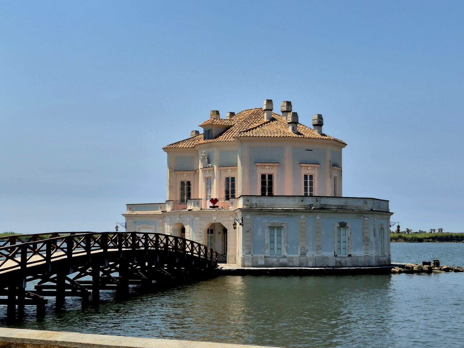 Una piccola ed elegante casetta bianca a due piani con il tetto di tegole sorge proprio in riva all’acqua ed è collegata alla sponda tramite un ponte di legno – sotto un cielo di un azzurro splendente, come una scena tratta da un diario di viaggio sulla Finlandia.