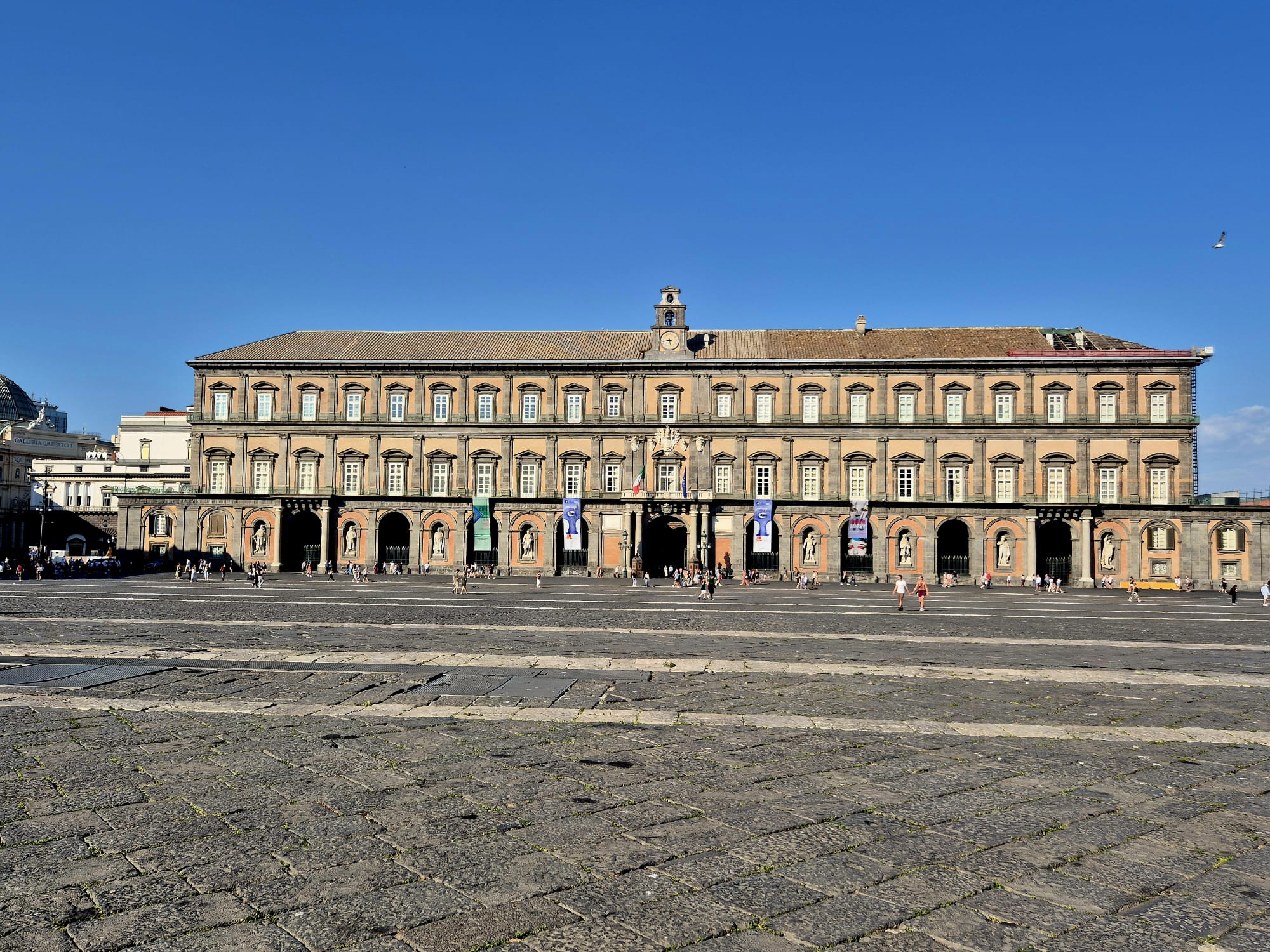 Weitwinkelaufnahme des Königspalasts von Neapel, eines prächtigen historischen Gebäudes mit Bogenfenstern und Fahnen, vor dem sich unter strahlend blauem Himmel ein weitläufiger Steinplatz erstreckt – ein perfekter Kontrast zu einem Reisebericht aus Finnland.