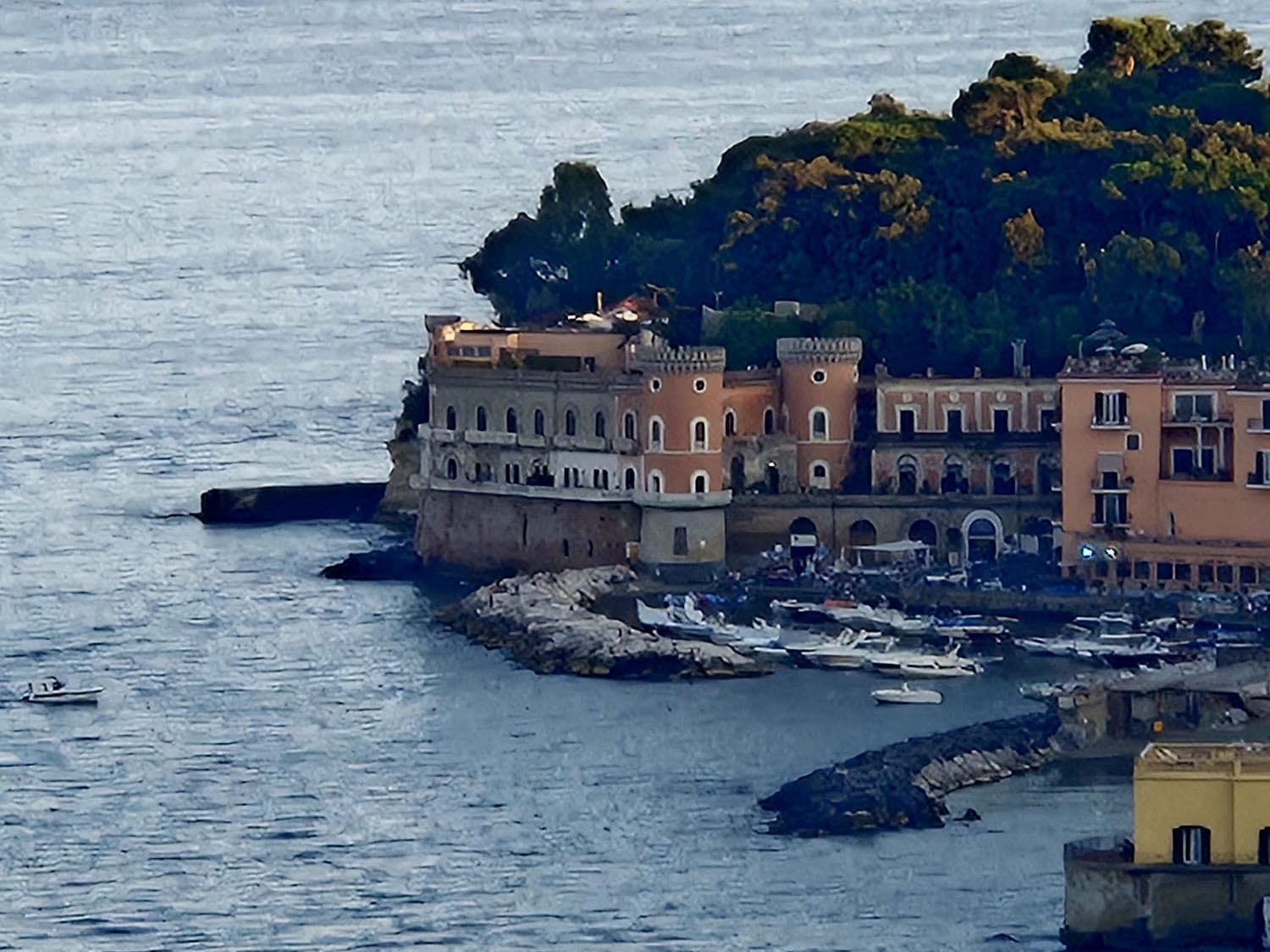 Un paesaggio costiero che ricorda un diario di viaggio dalla Finlandia, con edifici storici e un porto turistico pieno di barche sull'acqua tranquilla e blu, oltre ad alberi rigogliosi lungo una costa rocciosa.