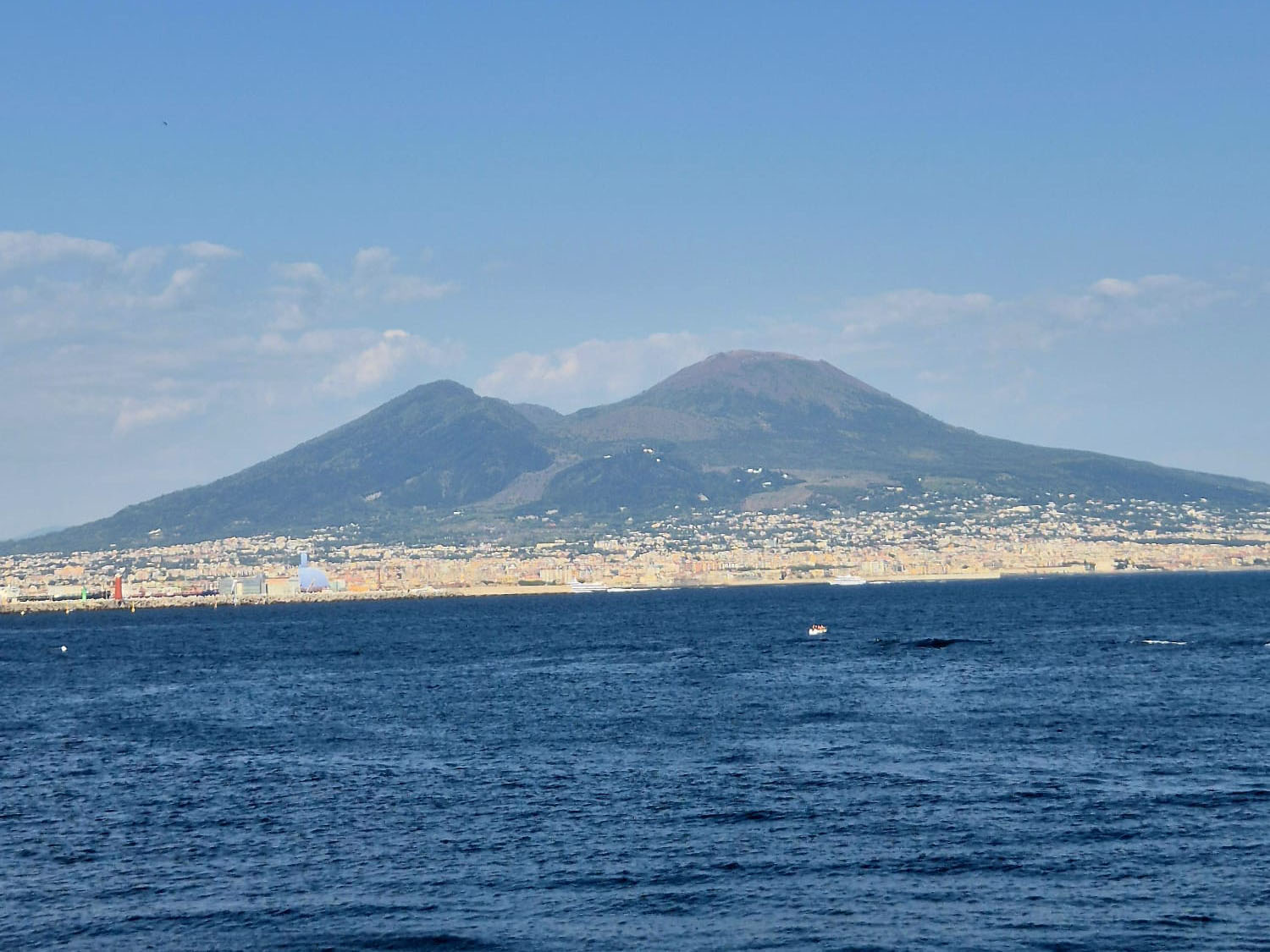 Una vista in lontananza di un grande vulcano a due cime che si erge sopra una città costiera – osservato attraverso un mare blu e calmo sotto un cielo limpido – ricorda le scene di un diario di viaggio sulla Finlandia.