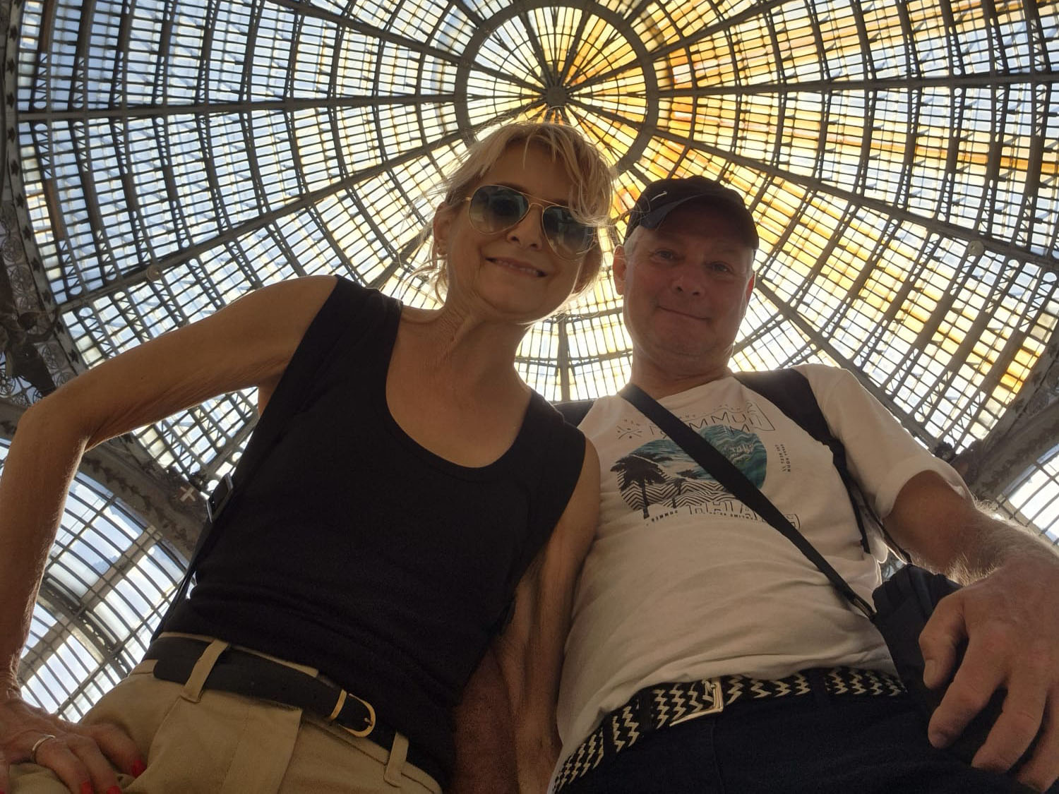 Una donna sorridente e un uomo stanno uno accanto all’altro e guardano verso la fotocamera sotto una grande cupola di vetro finemente decorata con un motivo geometrico illuminato dalla luce del sole, immortalando così un momento perfetto per il loro diario di viaggio in Finlandia.