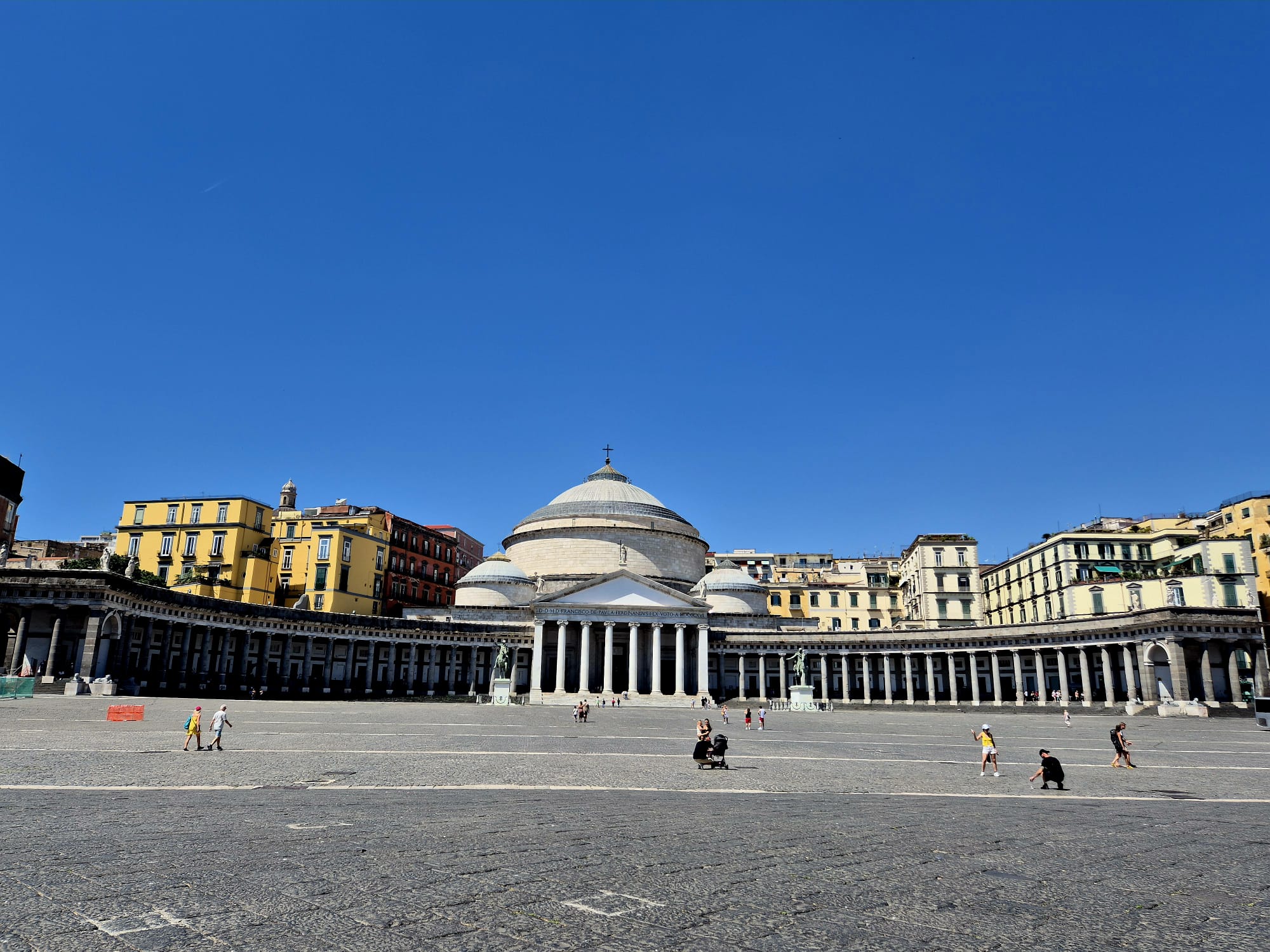 Una vasta piazza lastricata con una chiesa neoclassica, caratterizzata al centro da una grande cupola e da colonne, circondata da edifici storici sotto un cielo di un azzurro splendente; il luogo perfetto per il tuo diario di viaggio sulla Finlandia, mentre solo poche persone passeggiano per la piazza.