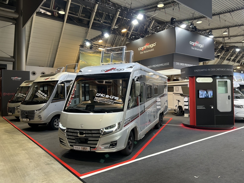 An einem Carthago-Messestand in der Halle sind zwei moderne Wohnmobile ausgestellt. Der Bereich ist mit Markenelementen, Informationsschildern und einem digitalen Kiosk ausgestattet; die Fahrzeuge werden unter heller Beleuchtung auf einem schwarzen Teppich präsentiert.