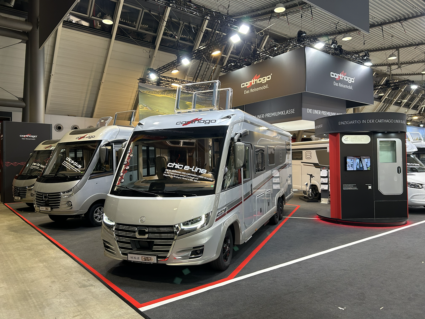 An einem Carthago-Messestand in der Halle sind zwei moderne Wohnmobile ausgestellt. Der Bereich ist mit Markenelementen, Informationsschildern und einem digitalen Kiosk ausgestattet; die Fahrzeuge werden unter heller Beleuchtung auf einem schwarzen Teppich präsentiert.