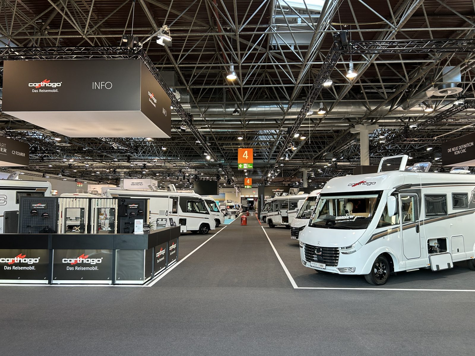 Ausstellungshalle mit mehreren ausgestellten weißen Reisemobilen, schwarzen Infoständen mit der Aufschrift „Carthago“ und Wegweisern an der Decke. Über dem geräumigen, mit Teppichboden ausgelegten Bereich sind Deckenleuchten und Metallträger zu sehen – dies vermittelt die Atmosphäre einer Carthago-Werksbesichtigung, bei der Besucher Premium-Reisemobile aus nächster Nähe erkunden können.